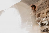 Mulher atraente com camisa quadriculada desabotoada andando perto de água do mar na costa rochosa contra montanhas no dia ensolarado no campo — Fotografia de Stock