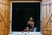 Два афроамериканских брата без рубашки смотрят в открытое окно деревянного дома, веселясь дома вместе — стоковое фото