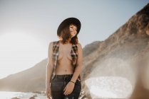 Mulher atraente com camisa quadriculada desabotoada andando perto de água do mar na costa rochosa contra montanhas no dia ensolarado no campo — Fotografia de Stock