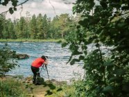 Vista posterior del hombre de mediana edad que se inclina y toma una foto de hermoso paisaje desde el otro lado del río - foto de stock