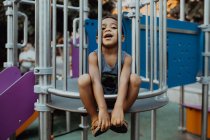 Afro americano ragazzo con divertente faccia guardando fotocamera mentre seduto dietro griglia su parco giochi in parco — Foto stock