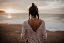 Vue arrière de la jeune femelle en tenue décontractée debout sur la plage de sable vers la mer orageuse pendant le coucher du soleil dans la nature — Photo de stock
