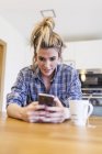 Schöne junge Frau, die zu Hause frühstückt und Spaß hat und mit dem Smartphone chattet — Stockfoto