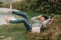 Молодий підліток з окулярами віртуальної реальності лежить на траві на відкритому повітрі біля озера з книгою і сміється вголос — стокове фото