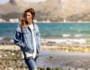Young female in denim outfit keeping hands in pockets and looking away while walking on coast near sea on windy day — Stock Photo