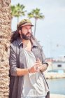 Стильный бородатый мужчина с длинными волосами, опирающийся на пальму — стоковое фото