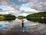 Vista posterior del hombre parado en el lago y abriendo los brazos mientras disfruta del paisaje con bosques y casas en Finlandia - foto de stock