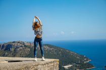 Красивая молодая женщина в повседневной одежде наслаждается солнцем, стоя на вершине цементного блока против безоблачного голубого неба возле моря — стоковое фото