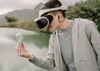 Молодой подросток, играющий в симуляцию виртуальной реальности с виртуальными очками, стоящими у озера, держа в руках световую лампочку и энергетическую концепцию — стоковое фото