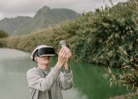 Young adolescent playing a virtual reality simulation with vr glasses standing near a lake holding a light bulb iinovation and energy concept — Stock Photo