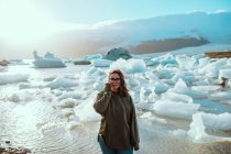 Bella giovane donna in abito casual guardando la fotocamera mentre in piedi sulla costa contro l'acqua increspata e lo scioglimento del ghiaccio nella soleggiata giornata primaverile — Foto stock
