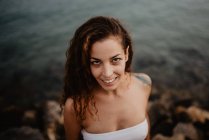 Vista lateral de uma jovem mulher bonita em roupa de banho sorrindo e olhando para a câmera enquanto está perto de água calma do mar na natureza — Fotografia de Stock