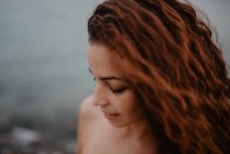 Vue latérale de jolie jeune femme en maillot de bain souriant et regardant vers le bas tout en se tenant près de l'eau calme de la mer dans la nature — Photo de stock