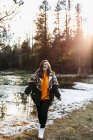 Young joyful female hiker on shore of small pond in snow among trees in sunny woods — Stock Photo