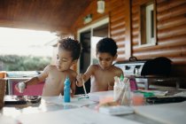 Due ragazzi afroamericani a torso nudo che usano vernice brillante per fare immagini astratte sul tavolo a casa — Foto stock