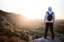 Back view of male hiker in sportswear standing on rocky cliff above terrain looking at picturesque landscape in morning sunlight — Stock Photo