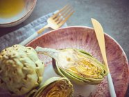 Whole and halved fresh artichokes on pink ceramic plate on table — Stock Photo