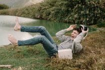 Jeune adolescent avec des lunettes de réalité virtuelle étendu sur l'herbe à l'extérieur près d'un lac avec un livre et riant à haute voix — Photo de stock
