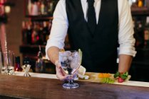 Crop anonyme jeune barman élégant travaillant derrière un comptoir de bar mélangeant boissons et fruits — Photo de stock