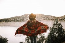 Vista trasera de la señora en sombrero y poncho mirando el cielo y las montañas al amanecer mientras está de pie en la orilla del tranquilo lago - foto de stock