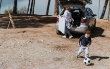 Little boy kicking ball while having fun near car and mother with canvas on sunny day in nature — Stock Photo