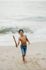 Divertente ragazzo afroamericano con bastone che gioca sulla riva sabbiosa vicino al mare — Foto stock