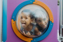 Dos pequeños chicos afroamericanos mirando a través de un ojo de buey y haciendo caras graciosas mientras jugaban juntos en el patio de recreo - foto de stock