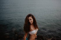 Вид збоку красива молода жінка в купальнику посміхається і дивиться на камеру, стоячи біля спокійної морської води в природі — стокове фото
