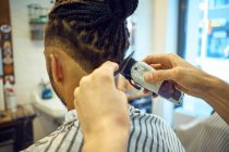 Vista de las cosechas por detrás del estilista anónimo haciendo un corte de pelo moderno con una navaja de afeitar a un cliente afroamericano sin rostro - foto de stock