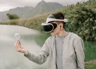 Jeune adolescent jouant une simulation de réalité virtuelle avec des lunettes vr debout près d'un lac tenant une ampoule iinovation et concept d'énergie — Photo de stock