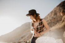Attraktive Frau mit aufgeknöpftem kariertem Hemd posiert in der Nähe von Meerwasser an felsiger Küste vor Bergen an sonnigen Tagen in der Natur — Stockfoto
