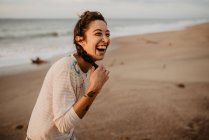 Jovem senhora em traje casual tocando trança e rindo em voz alta enquanto está em pé na praia arenosa — Fotografia de Stock