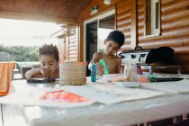 Dos chicos afroamericanos sin camisa usando pintura brillante para hacer cuadros abstractos en la mesa en casa - foto de stock