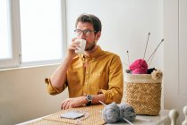 Homme adulte dégustant du thé chaud frais et regardant loin tout en étant assis à table près des aiguilles à tricoter et du fil — Photo de stock