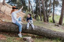 Lindo niño pequeño con pelota de fútbol mirando a la madre en gafas VR mientras está sentado en el tronco del árbol en el campo - foto de stock