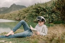 Giovane adolescente con occhiali di realtà virtuale posa su erba all'aperto vicino a un lago con un libro — Foto stock