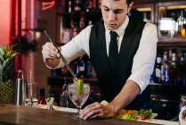 Jeune barman élégant travaillant derrière un comptoir de bar mélangeant boissons et fruits — Photo de stock