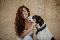 Funny dog licking cheek of excited young lady against weathered building wall on street — Stock Photo