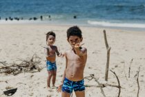 Афроамериканские дети смотрят в камеру и демонстрируют крошечную ракушку, стоя у синего моря — стоковое фото