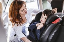 Smiling woman with dirty face fastening safety belt on cute boy in modern car — Stock Photo