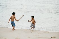 Dos divertidos hermanos afroamericanos con palos jugando juntos en la orilla arenosa cerca del mar - foto de stock