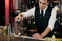 Joven barman elegante que trabaja detrás de un mostrador de bar verter bebida de coctelera a un vaso - foto de stock