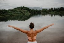 Vista trasera de una joven hembra con los brazos extendidos de pie cerca de un estanque tranquilo y disfrutando de la libertad en el campo - foto de stock
