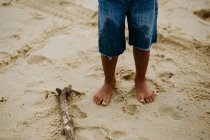 Ноги анонімного босоніж хлопчика в шортах, що стоять на мокрій піску біля дерев'яної палички, проводячи час на пляжі — стокове фото