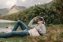 Jovem adolescente com óculos de realidade virtual deitado na grama ao ar livre perto de um lago com um livro — Fotografia de Stock