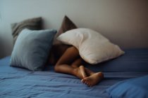 Criança descalça irreconhecível deitada sob almofadas em cama confortável no quarto acolhedor — Fotografia de Stock