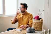Maschio adulto godendo di tè caldo fresco e guardando altrove mentre seduto a tavola vicino ferri da maglia e filati — Foto stock