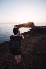 Back view of traveler with camera and backpack taking photo of rocky cliffs on seashore in sunset backlit — Stock Photo