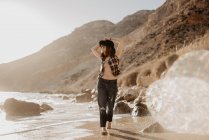 Attractive woman with unbuttoned checkered shirt walking near sea water on rocky coast against mountains on sunny day in countryside — Stock Photo