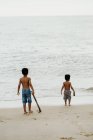Dos divertidos hermanos afroamericanos con palos jugando juntos en la orilla arenosa cerca del mar - foto de stock